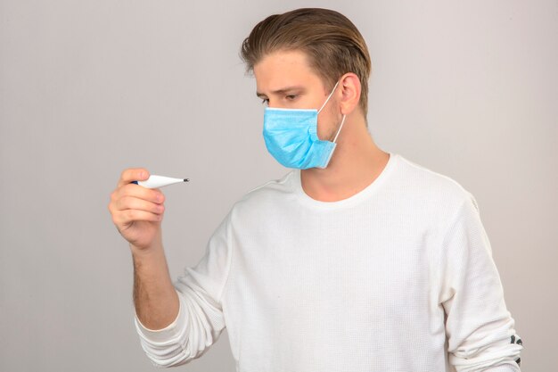 Junger kranker Mann in der medizinischen Schutzmaske, die digitales Thermometer über lokalisiertem weißem Hintergrund betrachtet