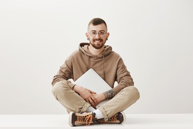 Junger kreativer Hipster-Student, der mit gekreuzten Beinen sitzt und Laptop hält