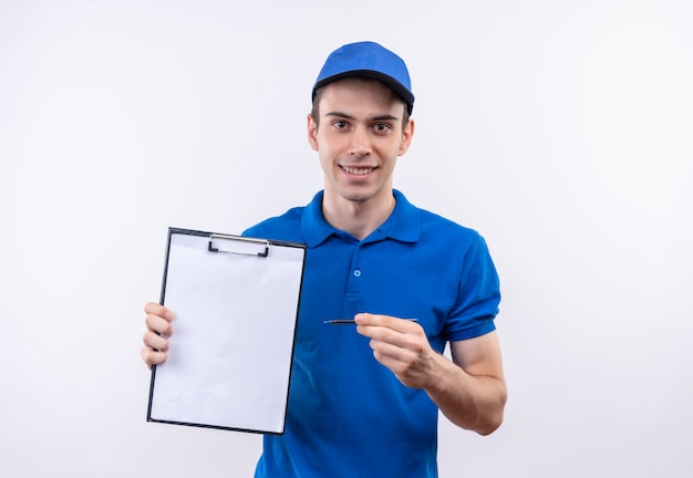 Junger Kurier, der blaue Uniform und blaue Kappe trägt, lächelt und zeigt auf Zwischenablage mit Stift