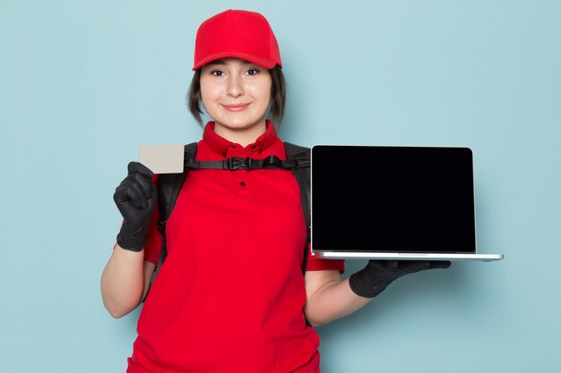 junger Kurier im roten Polo rote Kappe schwarze Handschuhe schwarzer Rucksack hält Laptop graue Karte auf blau
