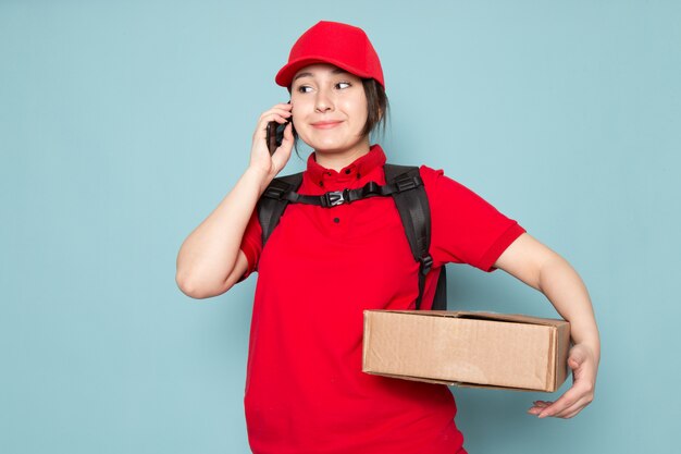 junger Kurier im roten Rucksack der roten Polo-roten Kappe, der Paket hält, das Telefon verwendet, das auf Blau lächelt