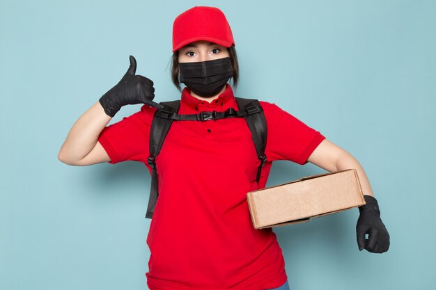 junger Kurier in roter Polo-roter Kappe schwarze sterile Schutzmaske schwarzer Rucksack, der Paket auf Blau hält