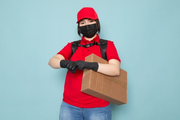 junger Kurier in roter Polo-roter Kappe schwarze sterile Schutzmaske schwarzer Rucksack, der Paket auf Blau hält