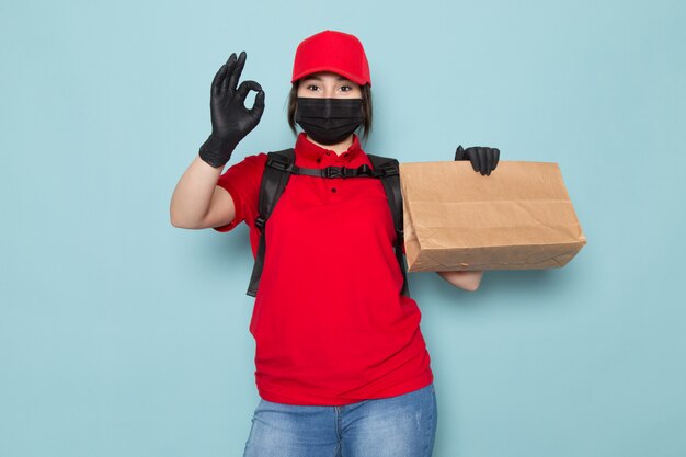 junger Kurier in roter Polo-roter Kappe schwarze sterile Schutzmaske schwarzer Rucksack, der Paket auf Blau hält