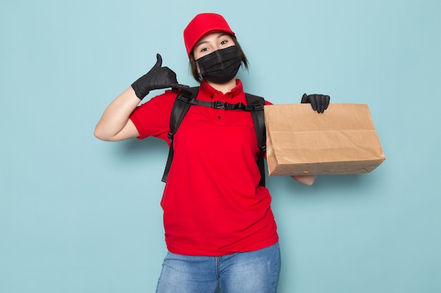 junger Kurier in roter Polo-roter Kappe schwarze sterile Schutzmaske schwarzer Rucksack, der Paket auf Blau hält