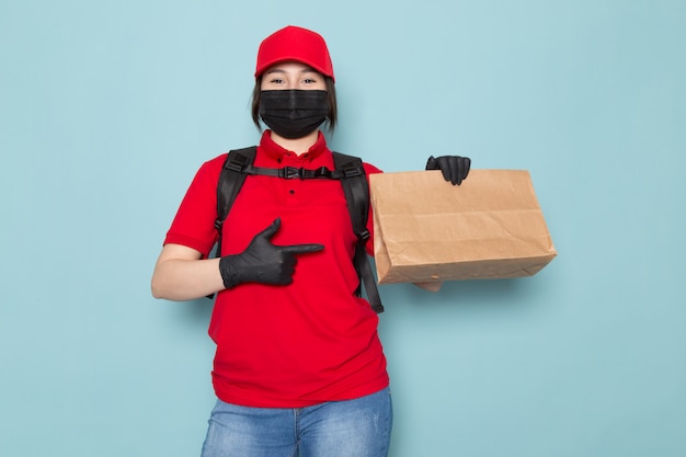 junger Kurier in roter Polo-roter Kappe schwarze sterile Schutzmaske schwarzer Rucksack, der Paket auf Blau hält