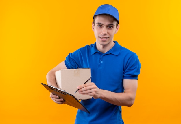 Junger Kurier mit blauer Uniform und blauer Mütze hält eine Schachtel und schreibt in die Zwischenablage