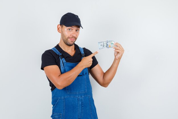 Junger Lader, der auf Dollarschein in Uniform zeigt und fröhlich, Vorderansicht schaut.