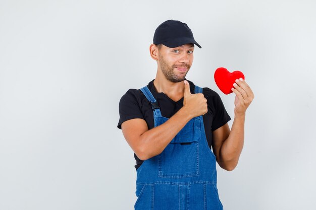 Junger Lader, der rotes Herz mit Daumen oben in Uniform hält und fröhlich schaut. Vorderansicht.