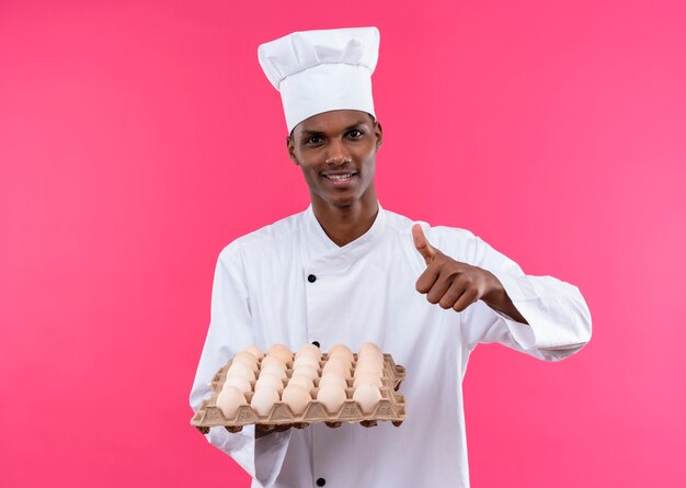Junger lächelnder afroamerikanischer Koch in der Kochuniform hält Charge von frischen Eiern und Daumen lokalisiert auf rosa Wand