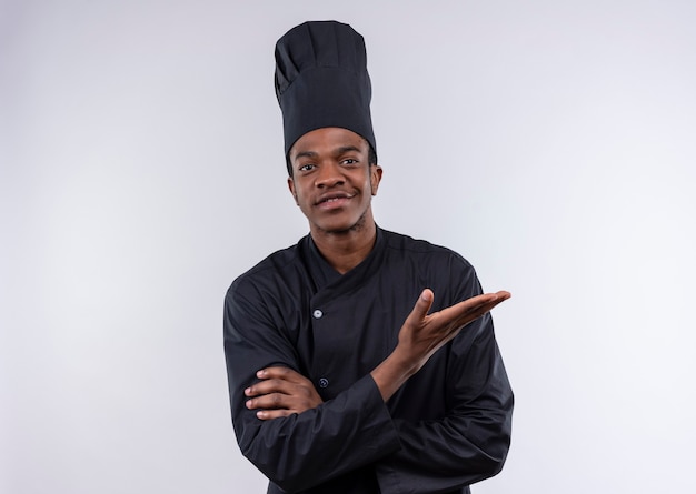 Junger lächelnder afroamerikanischer Koch in der Kochuniform kreuzt Arme und hält Hand offen lokalisiert auf weißer Wand
