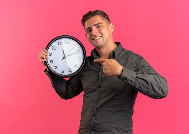 Junger lächelnder blonder hübscher Mann hält und zeigt auf Uhr lokalisiert auf rosa Raum mit Kopienraum