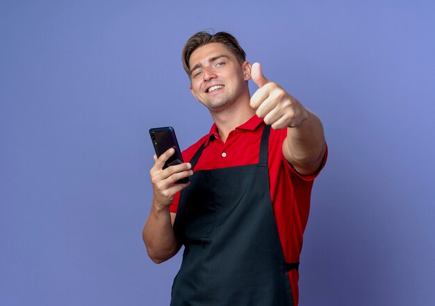 junger lächelnder blonder männlicher Friseur in Uniform hält Telefon Daumen hoch isoliert auf violettem Hintergrund