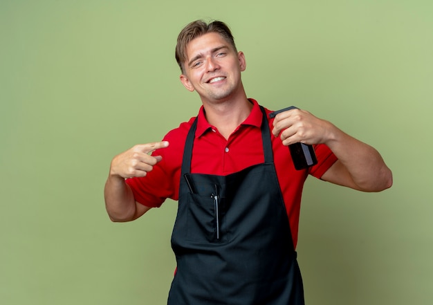 Kostenloses Foto junger lächelnder blonder männlicher friseur in uniform hält und zeigt auf sprühflasche lokalisiert auf olivgrünem raum mit kopienraum