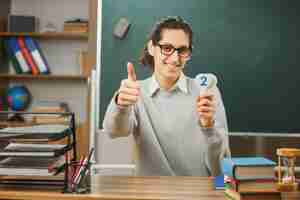 Kostenloses Foto junger lächelnder daumen hoch männlicher lehrer mit brille sitzt am schreibtisch und hält mathematikzahlen mit schulwerkzeugen im klassenzimmer