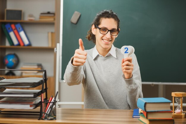 junger lächelnder Daumen hoch männlicher Lehrer mit Brille sitzt am Schreibtisch und hält Mathematikzahlen mit Schulwerkzeugen im Klassenzimmer