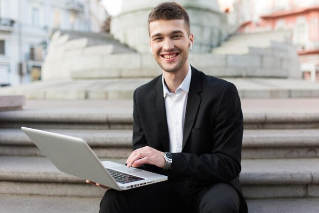 Junger lächelnder Geschäftsmann im klassischen schwarzen Anzug mit kabellosen Kopfhörern, der freudig in die Kamera schaut, während er den Laptop in den Händen hält und Zeit im Freien verbringt