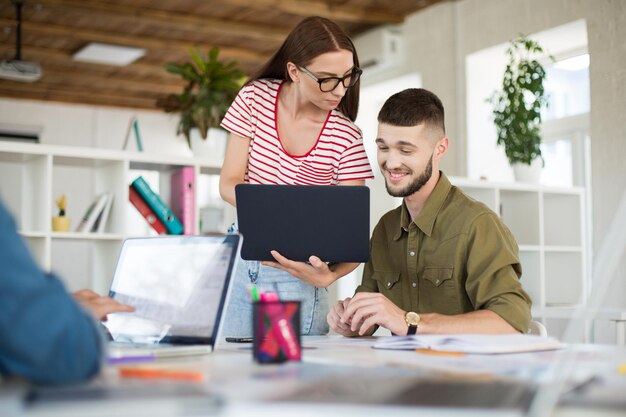 Junger lächelnder Geschäftsmann und Frau mit Laptop, die in einem modernen, gemütlichen Büro zusammenarbeiten Kreative Menschen verbringen Zeit bei der Arbeit
