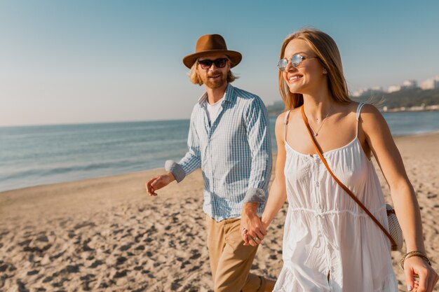 Junger lächelnder glücklicher Mann im Hut und in der blonden Frau, die zusammen auf Strand auf Sommerferienreisen laufen