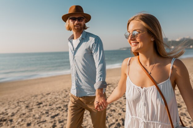 Junger lächelnder glücklicher Mann im Hut und in der blonden Frau, die zusammen auf Strand auf Sommerferienreisen laufen