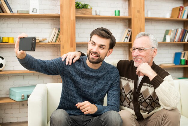 Junger lächelnder Kerl und gealterter Mann, der selfie auf Smartphone auf Sofa nimmt