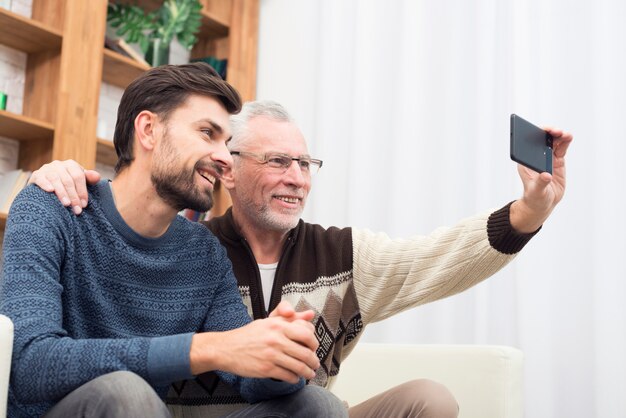 Junger lächelnder Kerl und gealterter netter Mann, der selfie am Handy auf Sofa nimmt