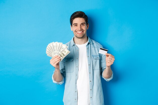 Junger lächelnder Mann, der Bargeld und Kreditkarte zeigt und auf blauem Hintergrund steht
