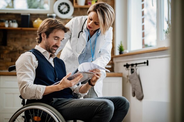 Junger lächelnder Mann im Rollstuhl und sein Arzt analysieren medizinische Berichte während des Hausbesuchs