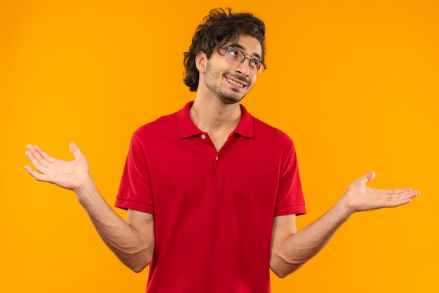 Junger lächelnder Mann im roten Hemd mit optischer Brille hält Hände offen lokalisiert auf orange Wand