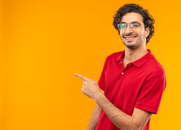 Junger lächelnder Mann im roten Hemd mit optischer Brille zeigt zur Seite und sieht auf orange Wand lokalisiert aus