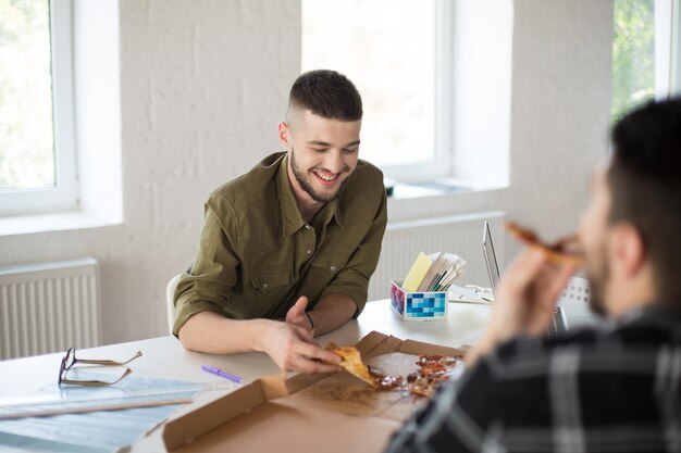 Junger lächelnder Mann mit Bart im Hemd, der glücklich Pizza isst, während er Zeit bei der Arbeit im modernen Büro verbringt