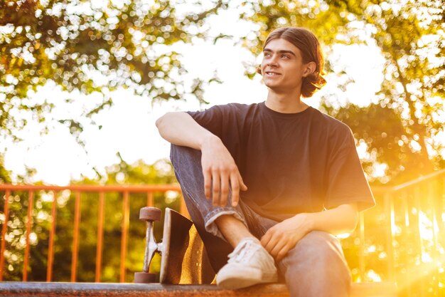 Junger lächelnder skater, der freudig mit skateboard im modernen skatepark mit schönem sonnenaufgang im hintergrund zur seite schaut