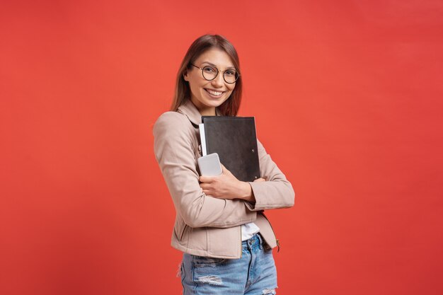 Junger lächelnder Student oder Praktikant in Brillen, die mit einem Ordner auf roter Wand stehen.