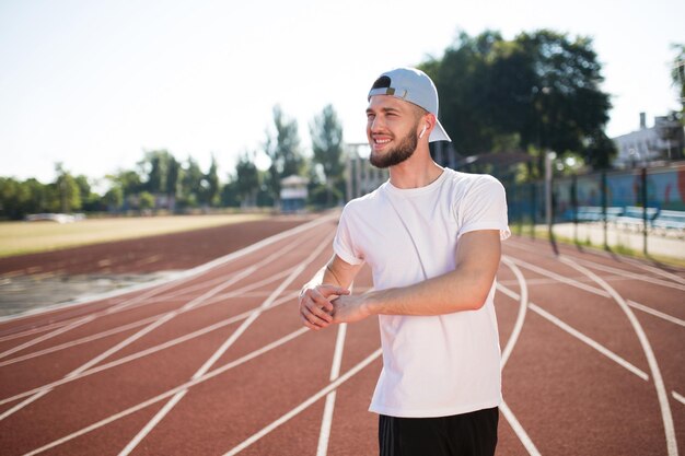 Junger lächelnder Typ mit drahtlosen Kopfhörern und weißer Kappe, der glücklich zur Seite schaut, während er Zeit auf dem Laufband des Stadions verbringt