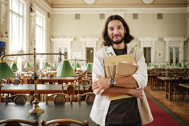 Junger lässig lächelnder männlicher Student mit Notizblöcken, der glücklich in die Kamera in der Bibliothek der Universität schaut