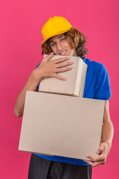 Junger Lieferbote, der blaues Poloshirt und gelbe Kappe trägt, die Pappkartons träumt, die Umarmungsboxen träumend lächelnd mit glücklichem Gesicht über lokalisiertem rosa Hintergrund träumt