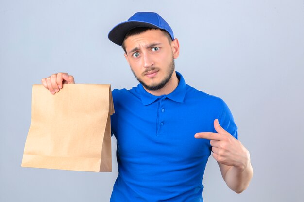 Junger Lieferbote, der blaues Poloshirt und Kappe hält, die Papierpaket mit Essen zum Mitnehmen zeigt, das auf dieses Paket mit Finger über lokalisiertem weißem Hintergrund zeigt