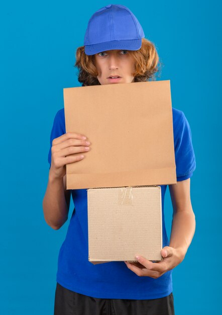 Junger Lieferbote, der blaues Poloshirt und Kappe hält, die Pappkarton und Papierpaket hält und überrascht steht über lokalisiertem blauem Hintergrund