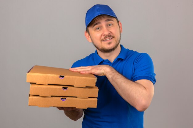 Junger Lieferbote, der blaues Poloshirt und Kappe hält, die Stapel von Pizzakästen betrachten, die Kamera freundlich über lokalisierten weißen Hintergrund lächelnd betrachten