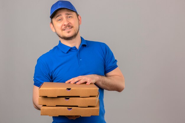 Junger Lieferbote, der blaues Poloshirt und Kappe hält, die Stapel von Pizzaschachteln betrachten Kamera mit freundlichem Lächeln über lokalisiertem weißem Hintergrund stehen