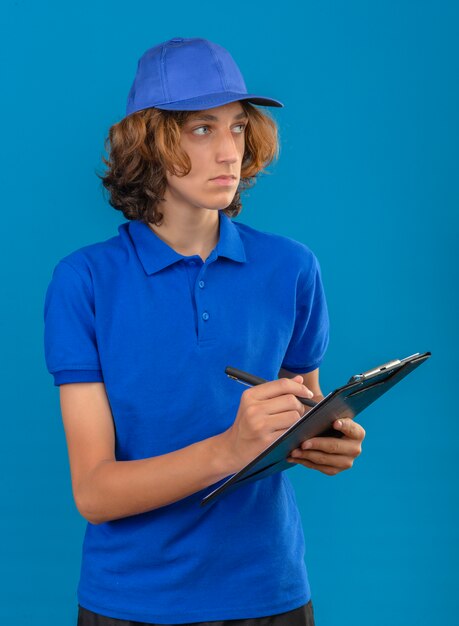Junger Lieferbote, der blaues Poloshirt und Kappe hält, die Zwischenablage hält, die etwas schaut, das beiseite schaut, das mit ernstem Gesicht über lokalisiertem blauem Hintergrund denkt