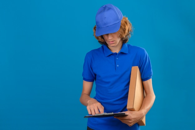 Kostenloses Foto junger lieferbote, der blaues poloshirt und kappe hält paket und zwischenablage mit leerzeichen lesen steht über lokalisiertem blauem hintergrund
