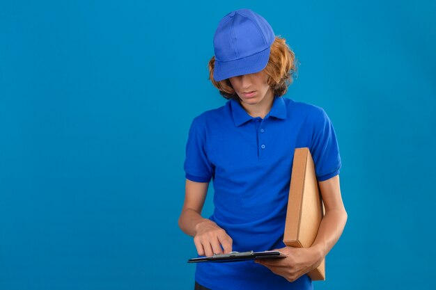 Junger Lieferbote, der blaues Poloshirt und Kappe hält Paket und Zwischenablage mit Leerzeichen lesen steht über lokalisiertem blauem Hintergrund