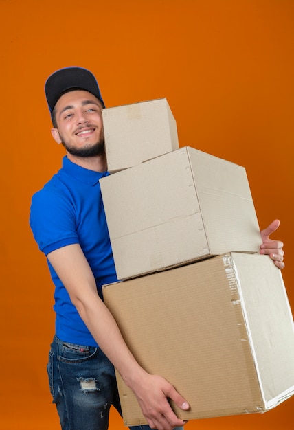 Junger Lieferbote, der blaues Poloshirt und Kappe trägt, die mit Stapel ob-Kästen stehen, die mit glücklichem Gesicht über lokalisiertem orangefarbenem Hintergrund lächeln