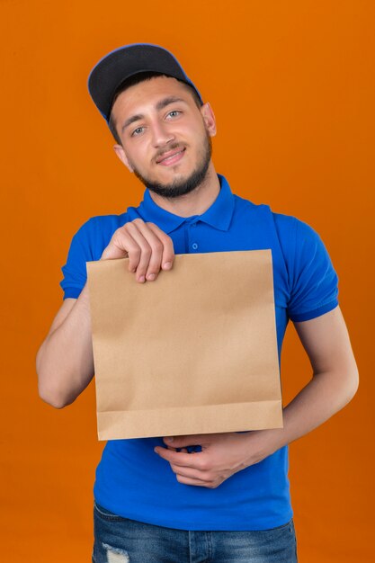 Junger Lieferbote, der blaues Poloshirt und Kappe zeigt Papierpaket betrachtet Kamera mit großem Lächeln auf Gesicht über lokalisiertem orange Hintergrund