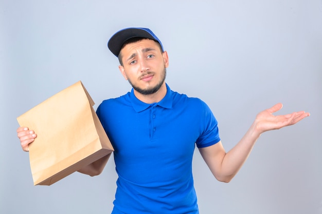 Junger Lieferbote, der blaues Poloshirt und Mütze trägt, die mit Papierpaket mit Essen zum Mitnehmen stehen, verwechselt mit Händen, die über lokalisiertem weißem Hintergrund erhoben werden