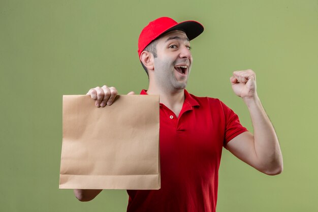 Junger Lieferbote, der rote Uniform hält, die Papierpaket hebt, das Faust feiert, das Sieg mit glücklichem Gesicht über lokalisiertem grünem Hintergrund feiert