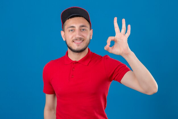 Junger Lieferbote, der rotes Poloshirt und Kappe trägt, die Kamera mit Lächeln tut, das ok Zeichen über lokalisiertem blauem Hintergrund tut