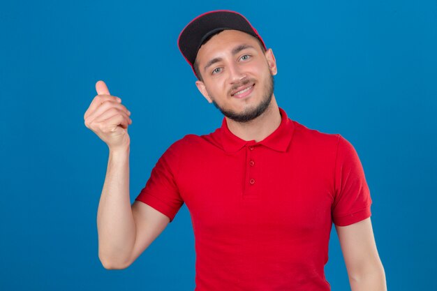 Junger Lieferbote, der rotes Poloshirt und Kappe trägt, die mit Lächeln auf Gesicht stehen und eine Geldgeste über lokalisiertem blauem Hintergrund tun