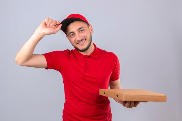 Junger Lieferbote, der rotes Poloshirt und Kappe trägt, die mit Stapel von Pizzaschachteln stehen, die Grußgestenberührungskappe freundlich über lokalisierten weißen Hintergrund lächelnd machen